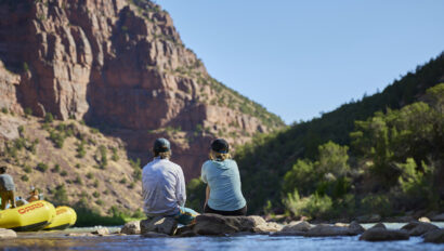 A couple sitting on a rock near a river gazing off at a towering red rock canyon wall