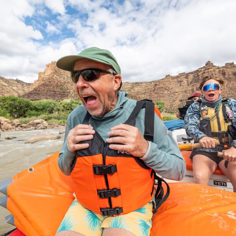 A man sits in the front of a raft with a big grin on his face as his expressive guide rows