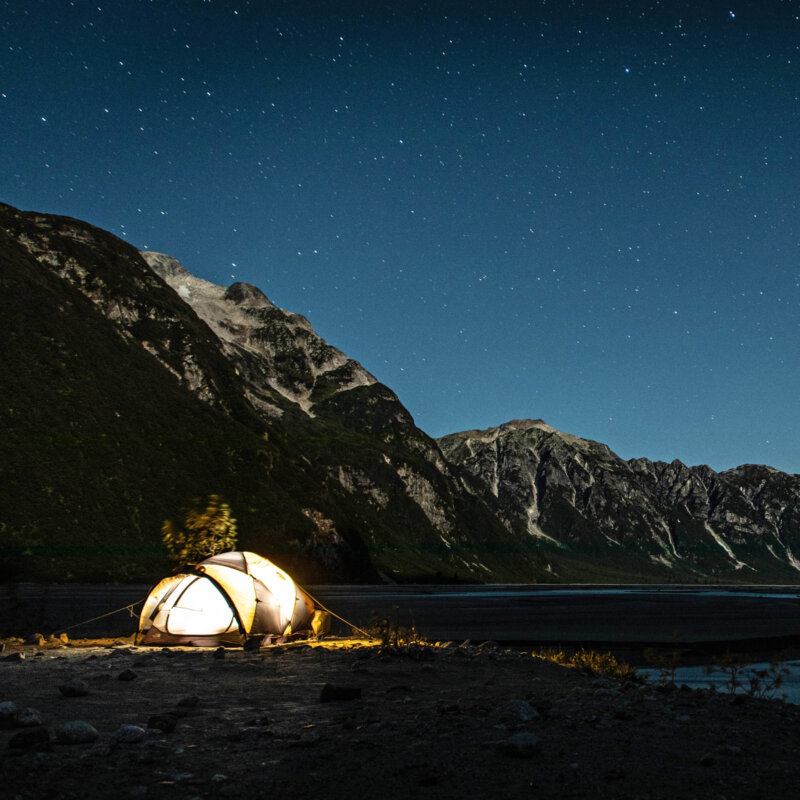 A tent lit from inside set up at camp with the the river and towering mountains behind it, and a twinkling sky above.