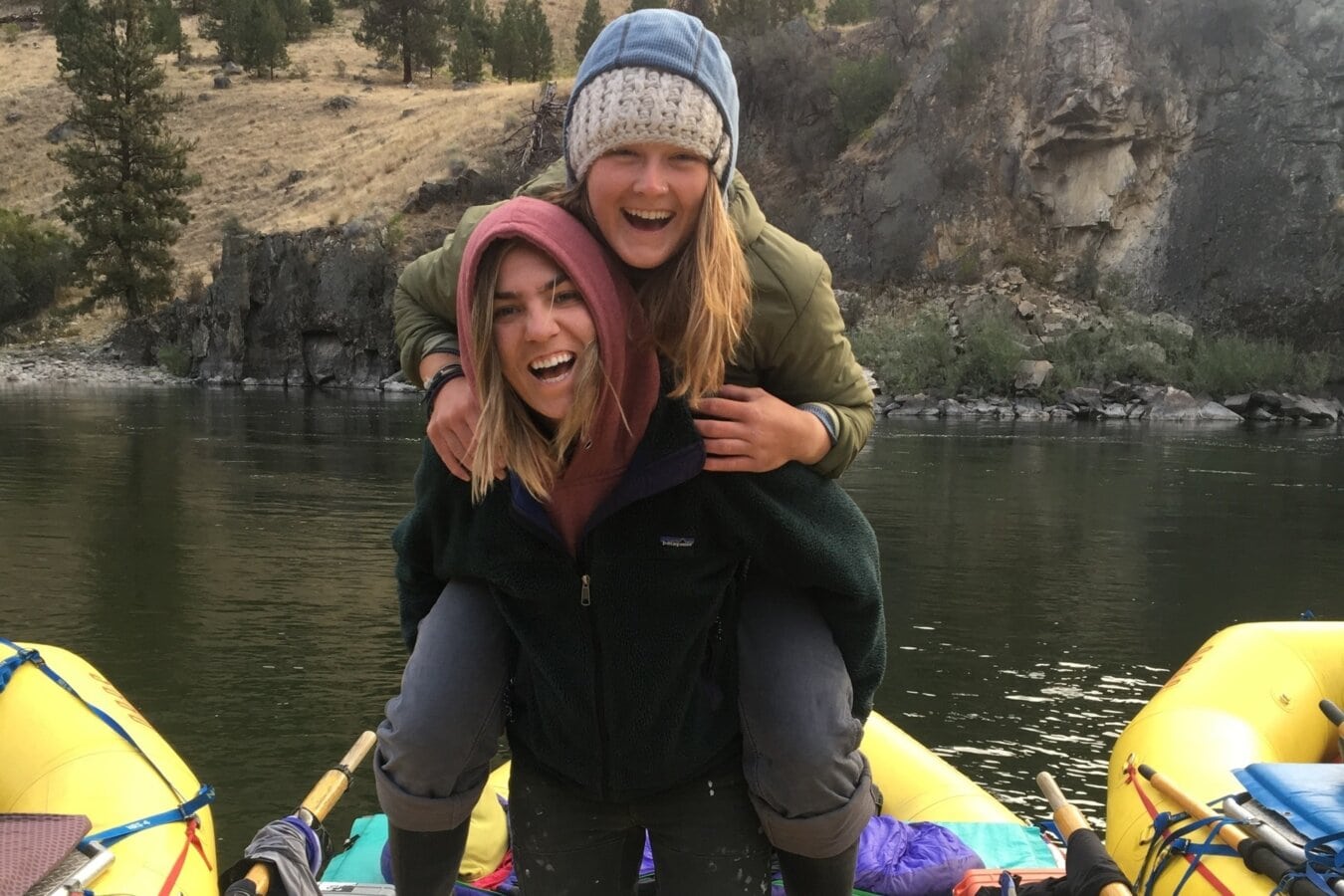 Two women laugh as they pose for the camera while one is giving the other a piggy back on a raft.