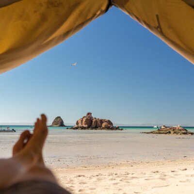 A serene beach view from inside a tent, showcasing turquoise waters, sandy shores, and rock formations under a clear blue sky.