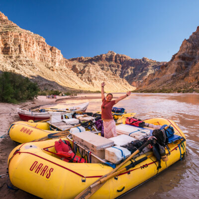 A guide stands on her raft and smiles at the camera with her hands in the air along the banks of the Colorado River.