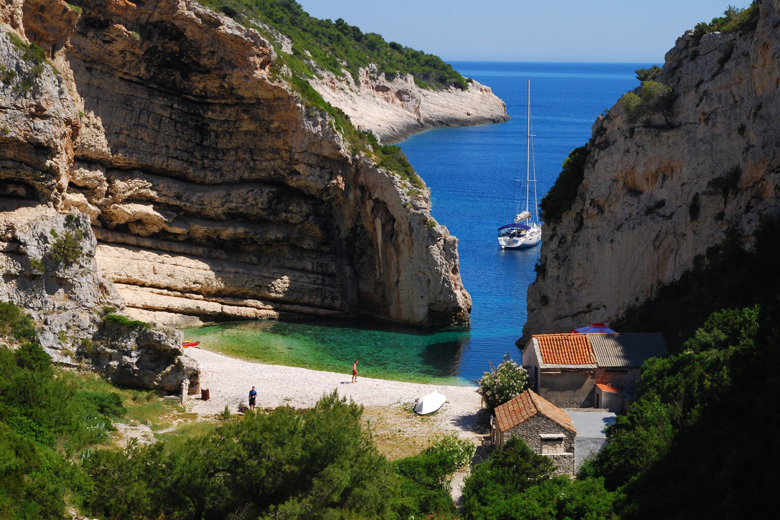 View from above of a cove with a sandy beach and small buildings and a sail boat sits offshore in Croatia.