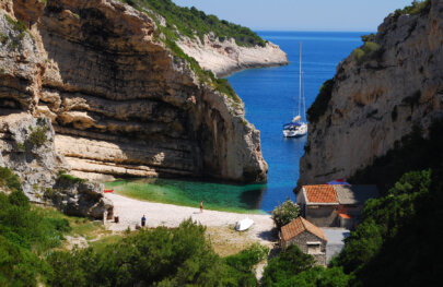 View from above of a cove with a sandy beach and small buildings and a sail boat sits offshore in Croatia.