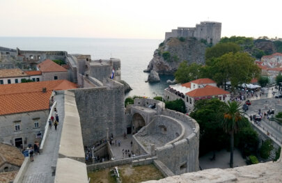 View of one of the charming sea-side town in Croatia as people walk along it's streets with a castle perched high on a hill in the background.