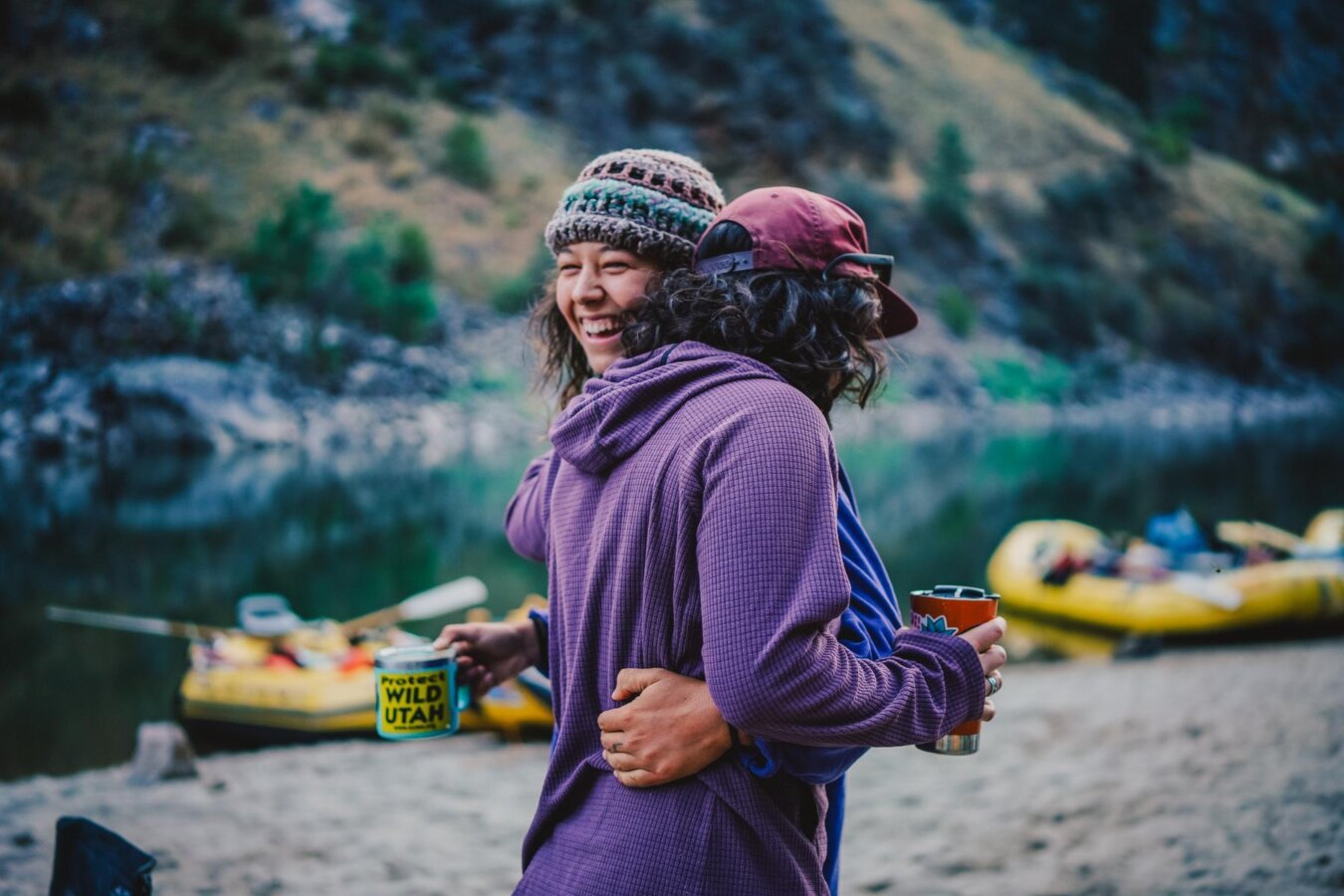 Two women hug and smile while holding coffee mugs on a sandy beach next to the Main Salmon River in Idaho.