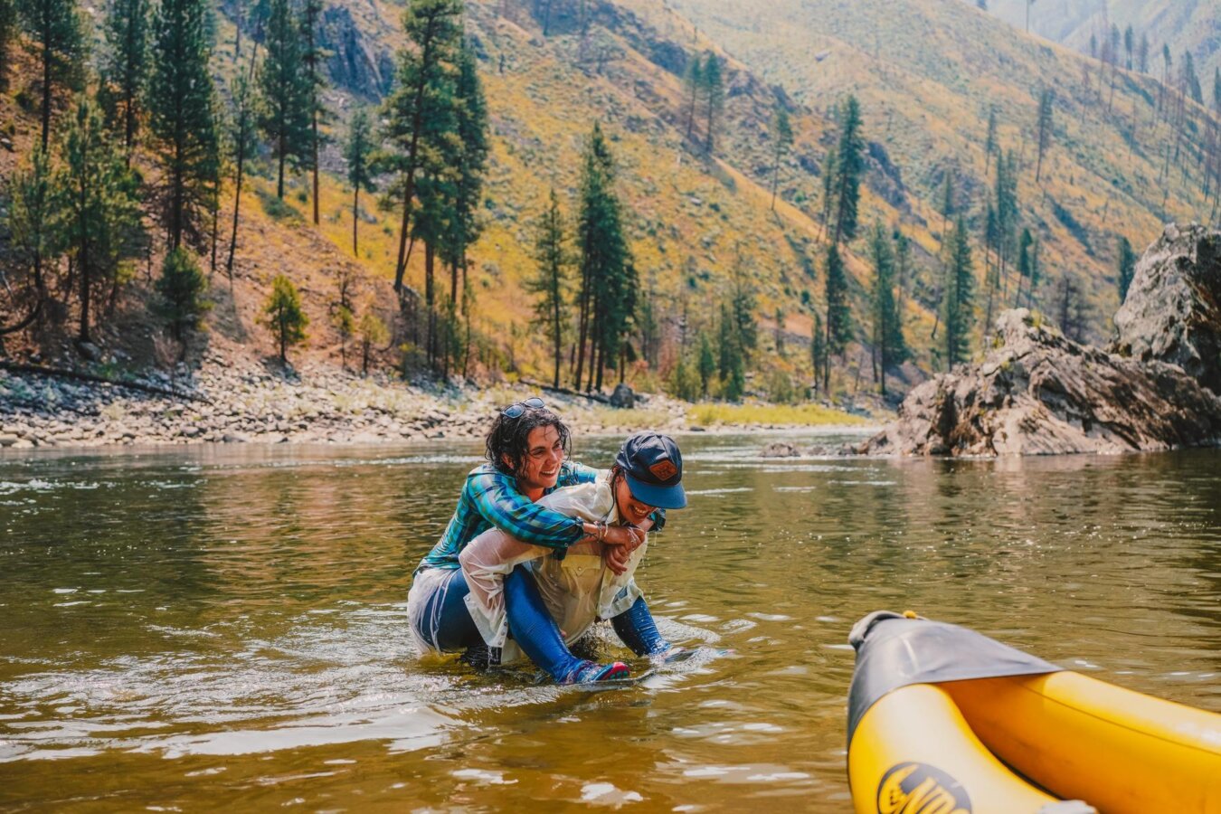A woman gives another woman a piggy back while walking in the river. Both are smiling.