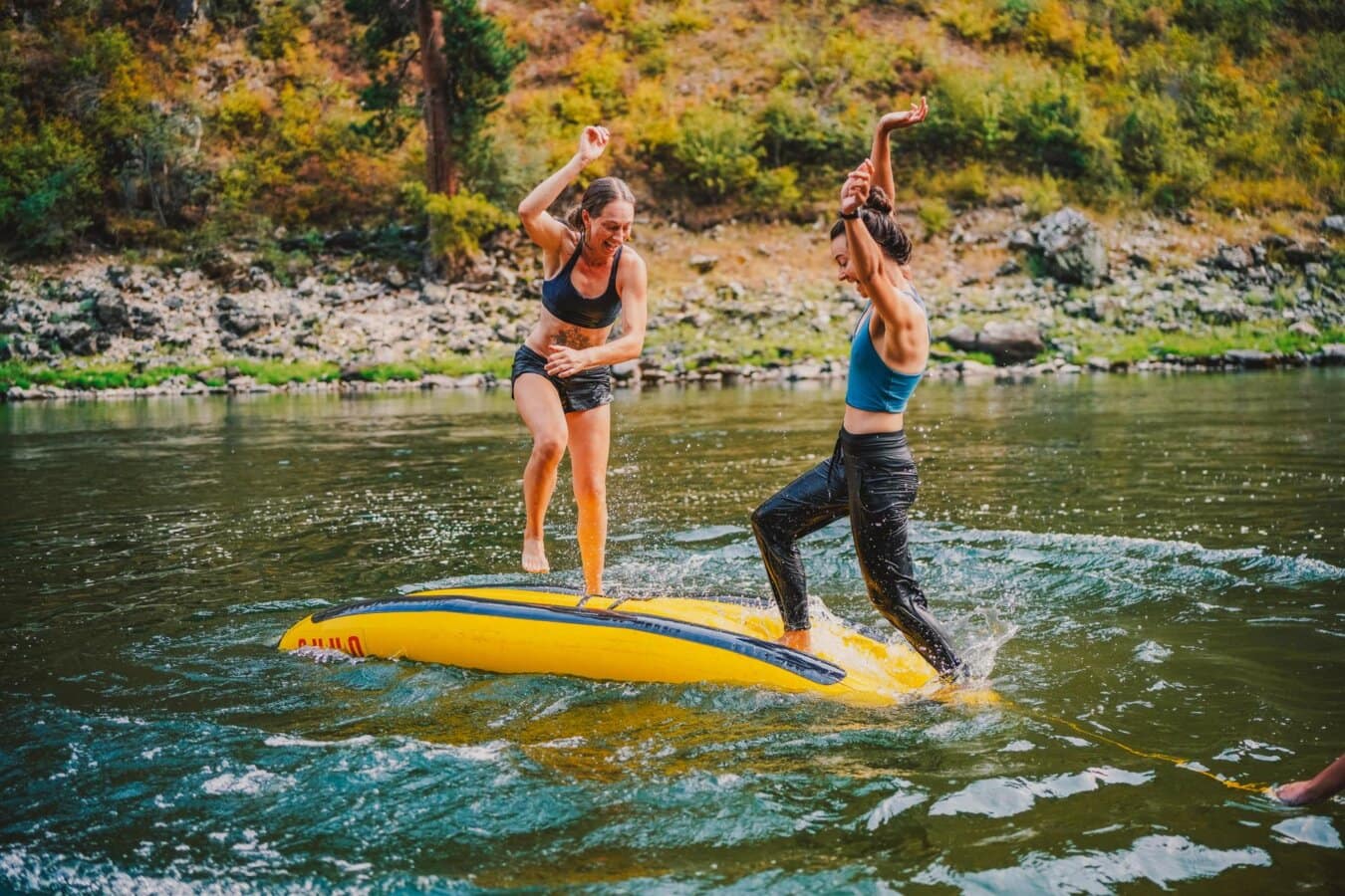 Two women play a balance game on a upside-down inflatable kayak floating in the Main Salmon River