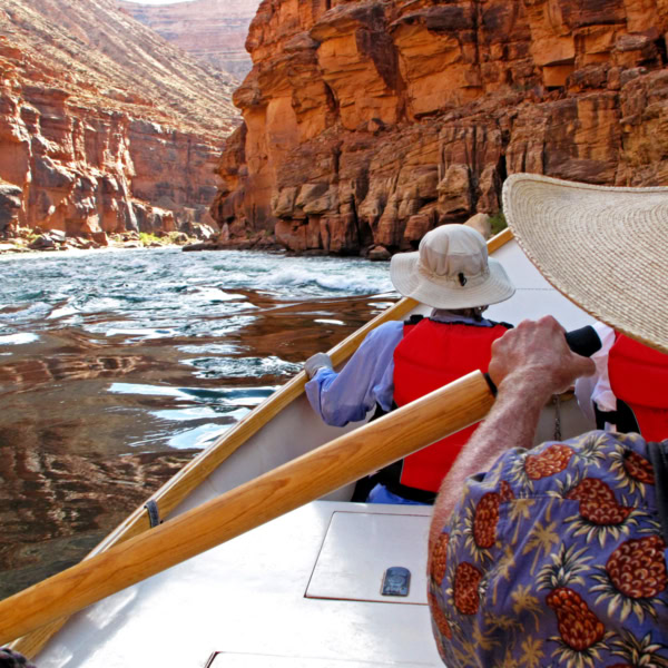 View downstream looking over OARS dory guide's shoulder with guest in PFD and sun hat hanging on in Grand Canyon