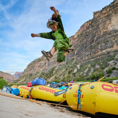 A kid wearing a dinosaur onsie striking a superhero pose mid-air as he jumps off the front of a yellow raft onto the sandy beach at camp.
