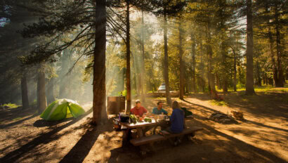 Three people sitting at a picnic table preparing food at a forested car camping campsite