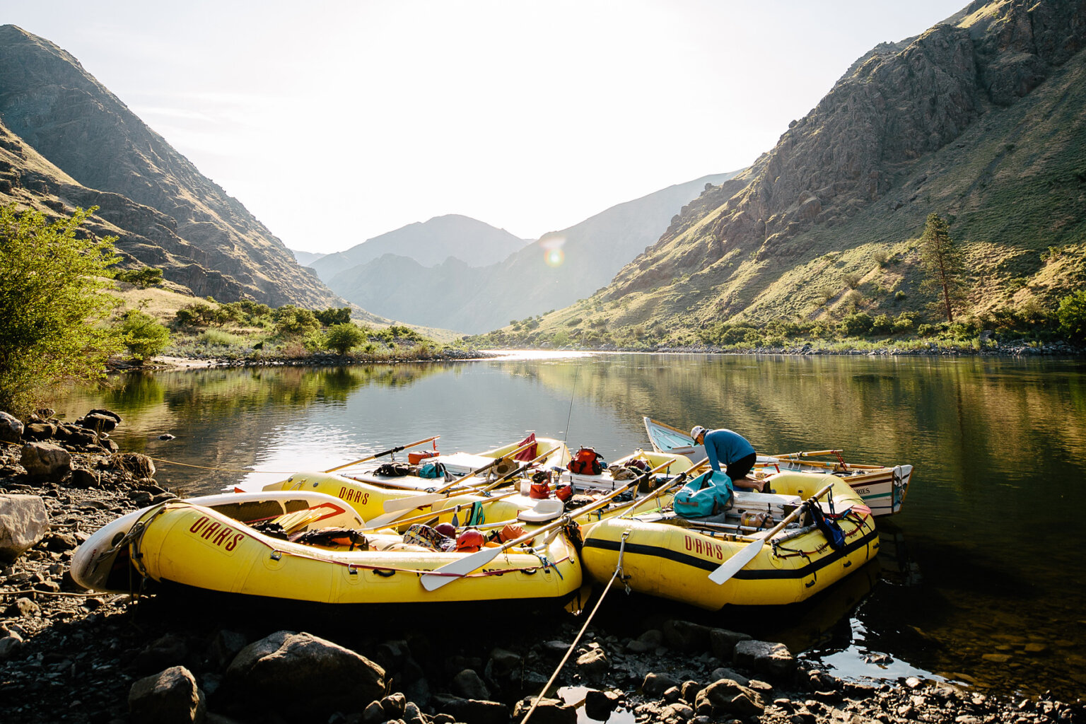 Several yellow rafts and a dory are tied up on the shoreline of the Snake River with rocky green hills rising all around and a guide works on one of the rafts.
