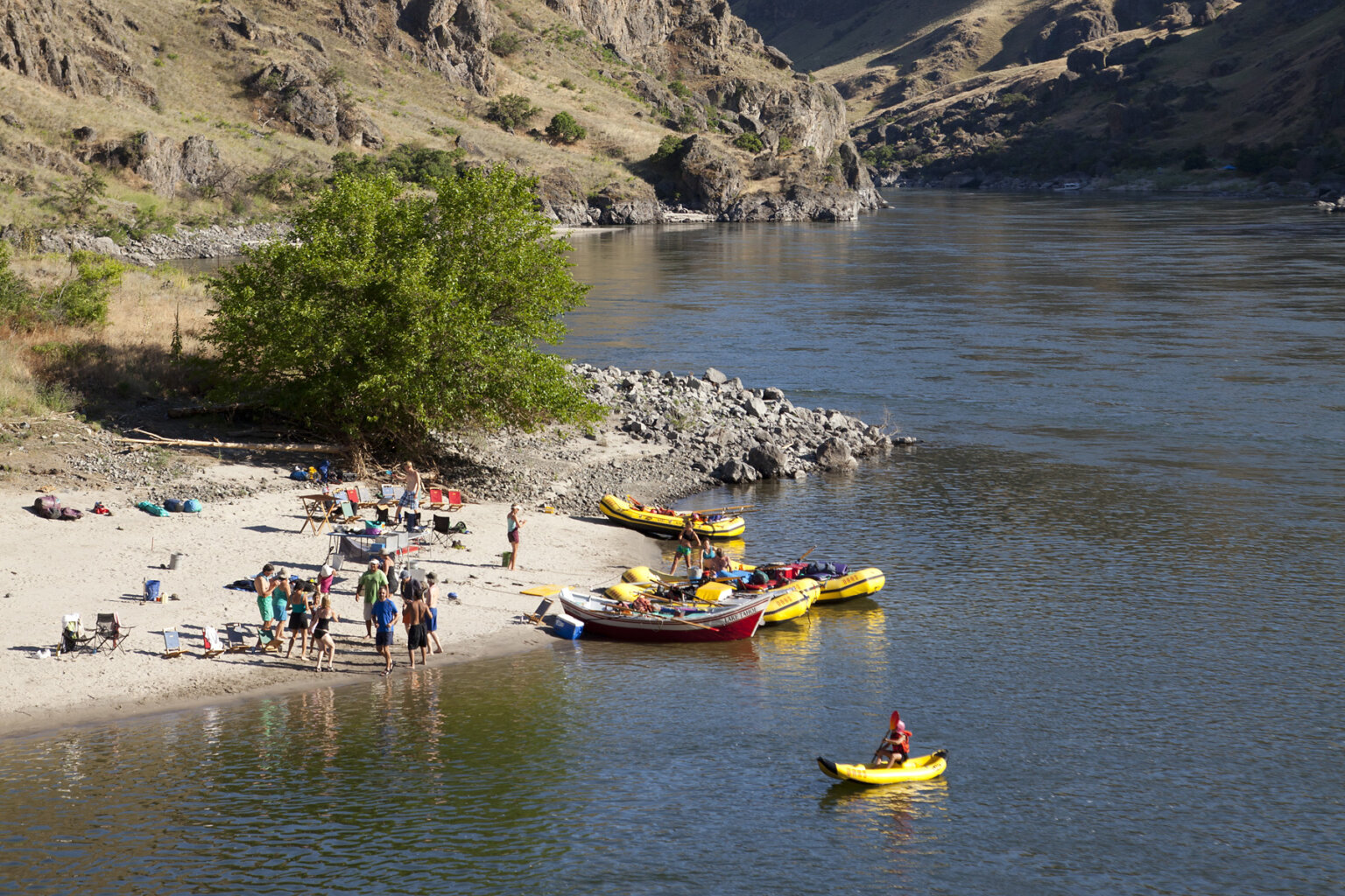 Shot of camp from high above; several guests play on the sandy beach and one guest is using a yellow inflatable kayak on the river close to shore.