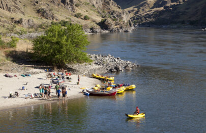 Shot of camp from high above; several guests play on the sandy beach and one guest is using a yellow inflatable kayak on the river close to shore.