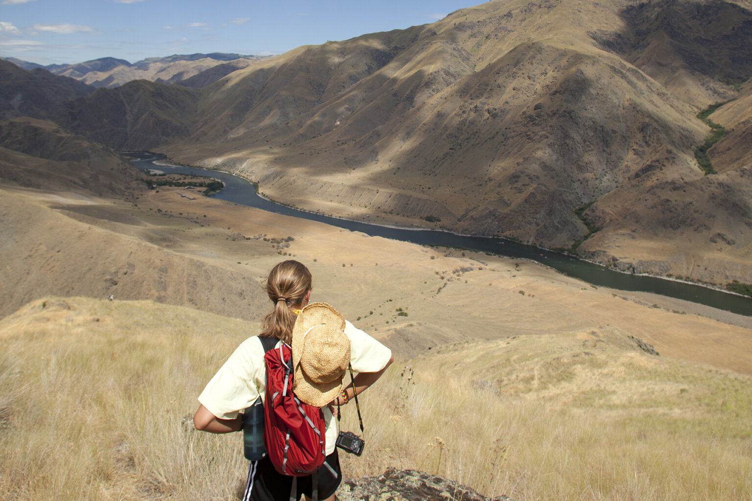 View from behind of a hiker taking in the view of the river below surrounded by golden rolling hillsides and mountains.