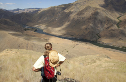 View from behind of a hiker taking in the view of the river below surrounded by golden rolling hillsides and mountains.