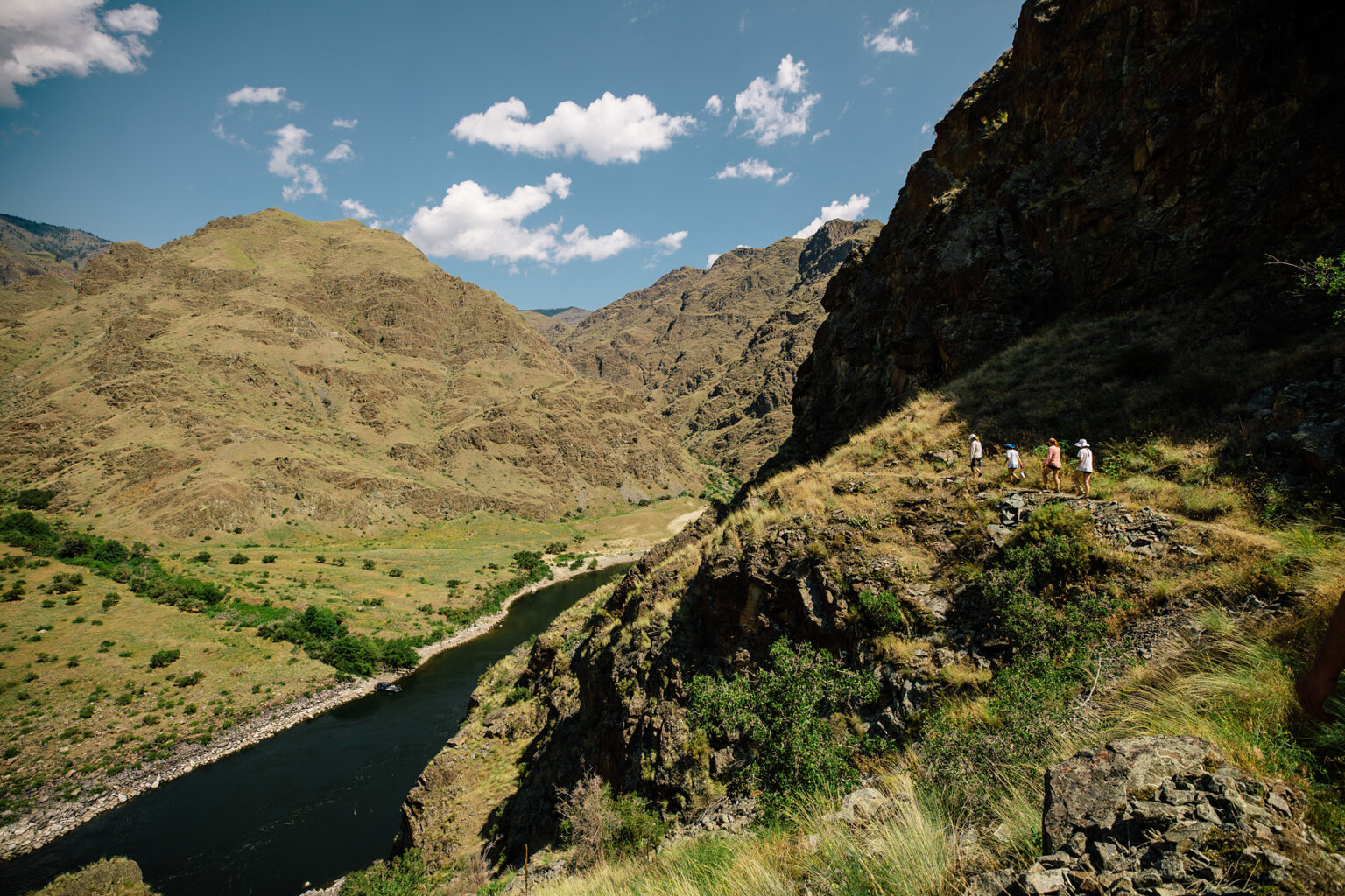 Four guests are seen in the distance walking along a trail high above the Snake River and verdant and golden rocky mountains rise all around.