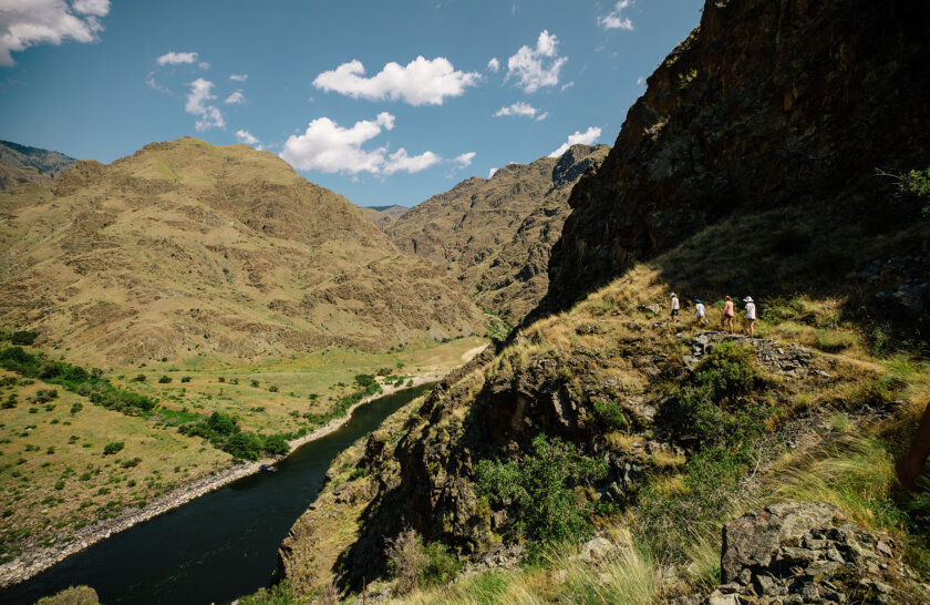 Four guests are seen in the distance walking along a trail high above the Snake River and verdant and golden rocky mountains rise all around.