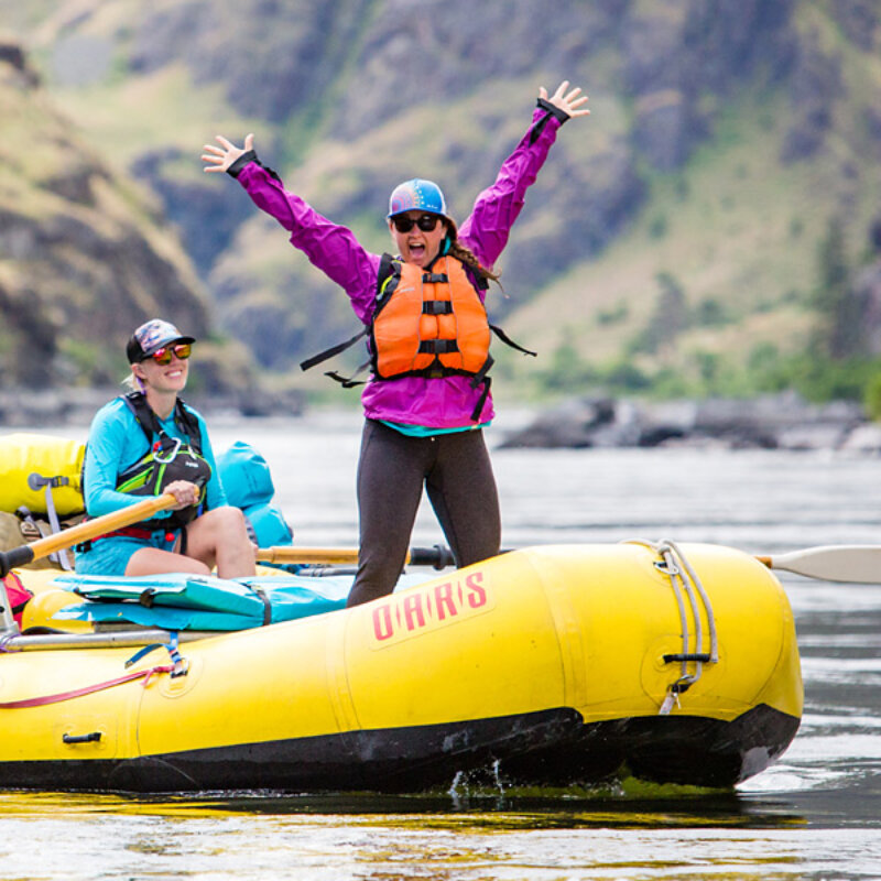 An excited woman stands in the front of a yellow raft with her hands in the air while a guide rows down the Snake River in Hells Canyon.