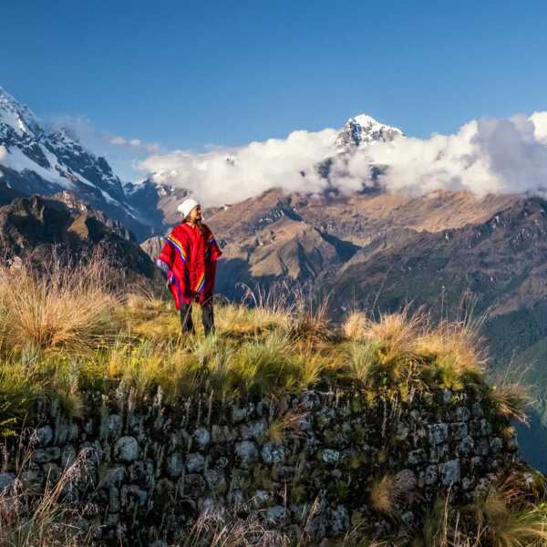A woman in a Peruvian poncho stands on a vista overlooking forrested and snow-capped mountains.