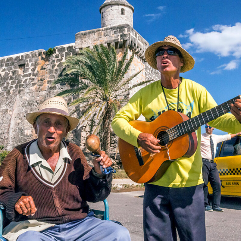 A Cuban man stands playing a guitar while an older Cuban man in a wheelchair plays maracas and they sing.
