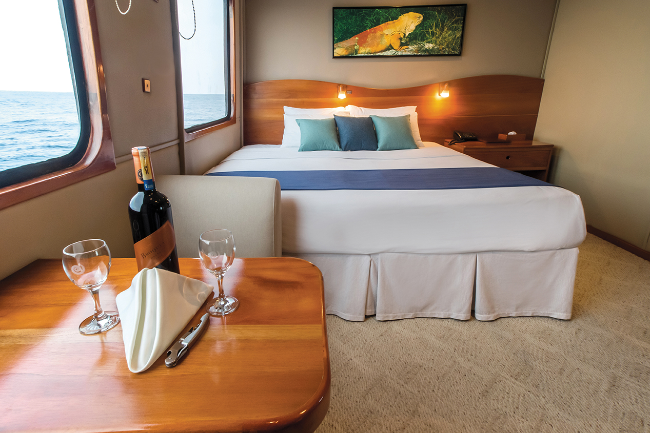 A bottle of wine and two glasses sit on a small table near the king bed of a suite on the Integrity yacht.