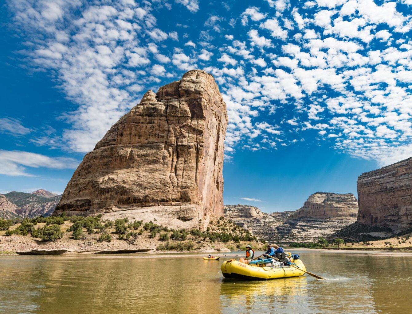 Yellow raft and inflatable kayak in front of a mountain of a boulder jutting out of the ground across the river.