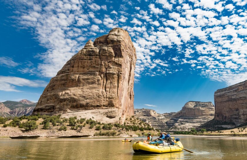 Yellow raft and inflatable kayak in front of a mountain of a boulder jutting out of the ground across the river.