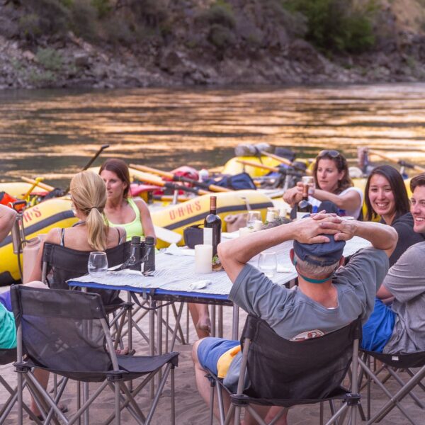 A group of adults sit around a linen draped table on a sandy beach drinking wine and socializing on an OARS Wine on the Salmon River trip.