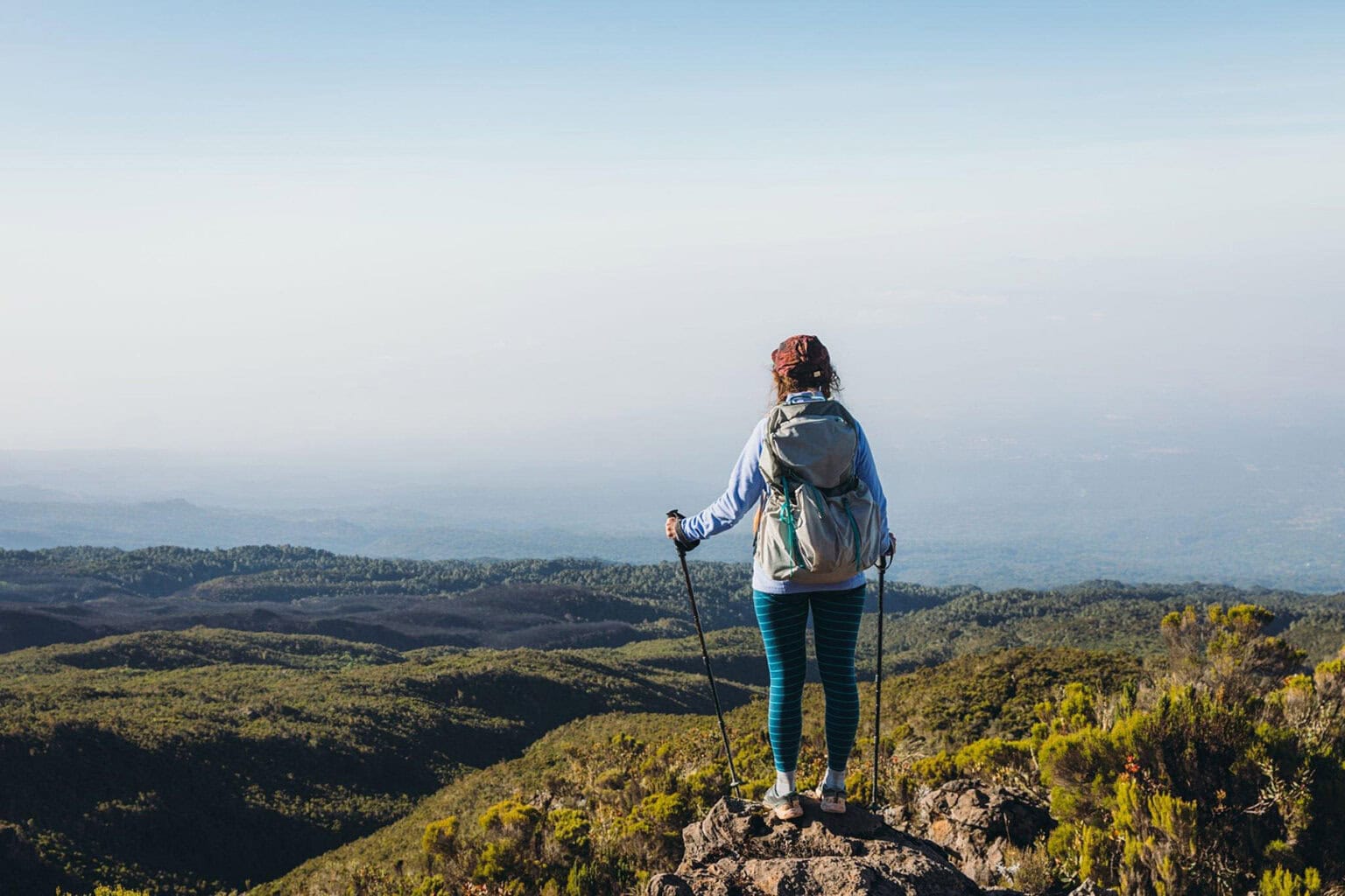 A hiker stands on a rocky ledge, gazing at a vast landscape of green hills under a clear sky, holding trekking poles.