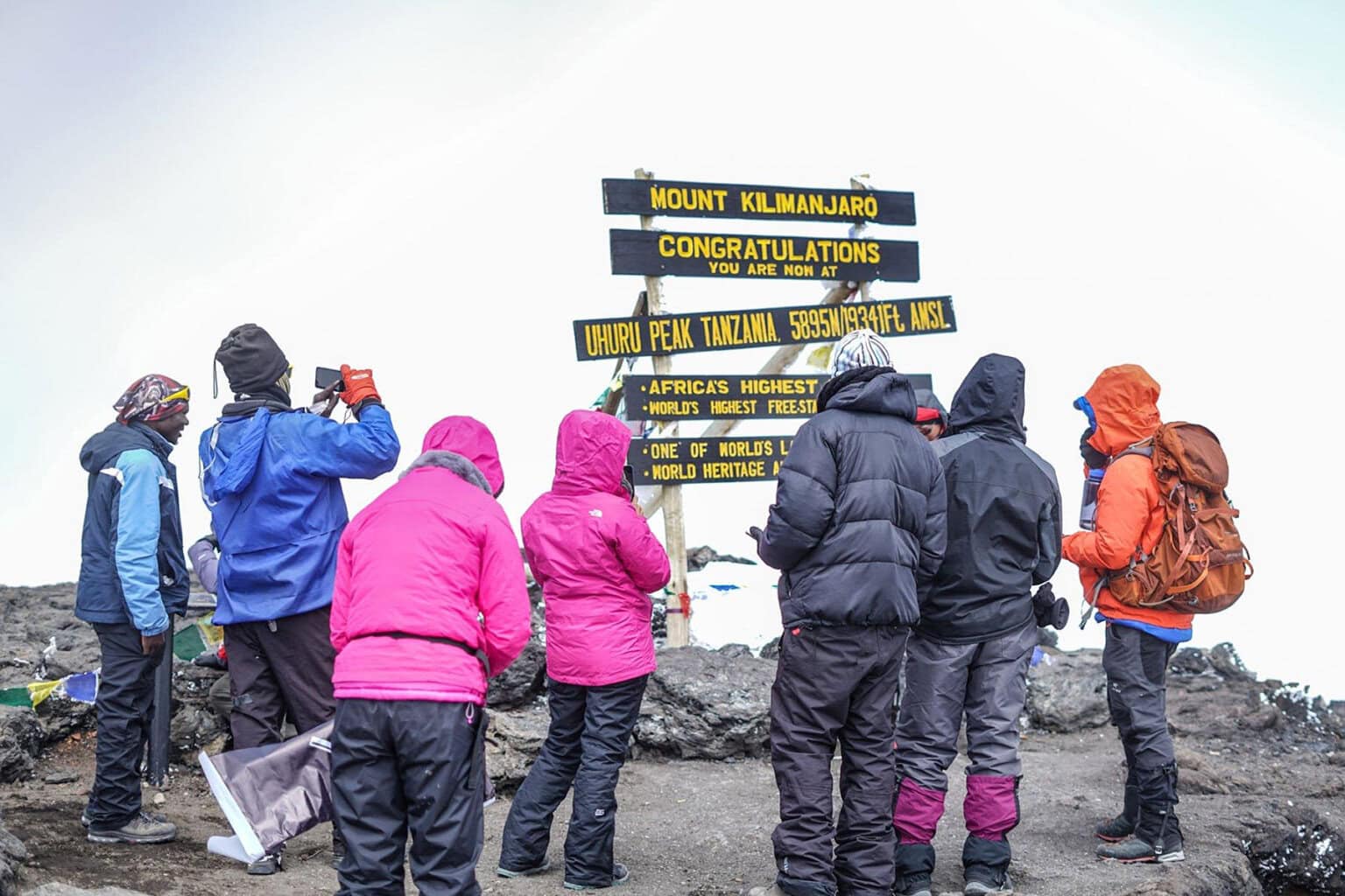 A group of climbers in colorful jackets stands at the summit of Mount Kilimanjaro, celebrating and taking photos near the iconic sign.