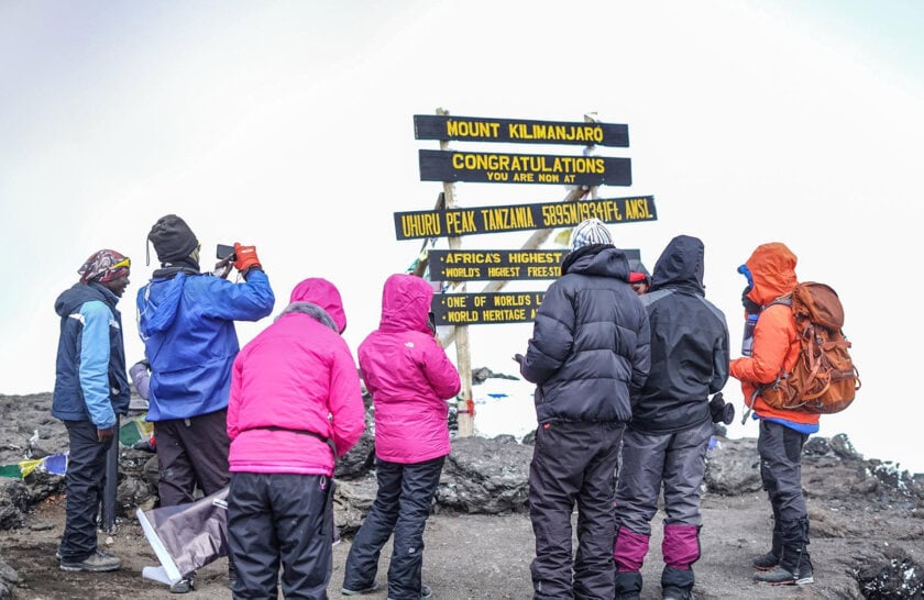 A group of climbers in colorful jackets stands at the summit of Mount Kilimanjaro, celebrating and taking photos near the iconic sign.