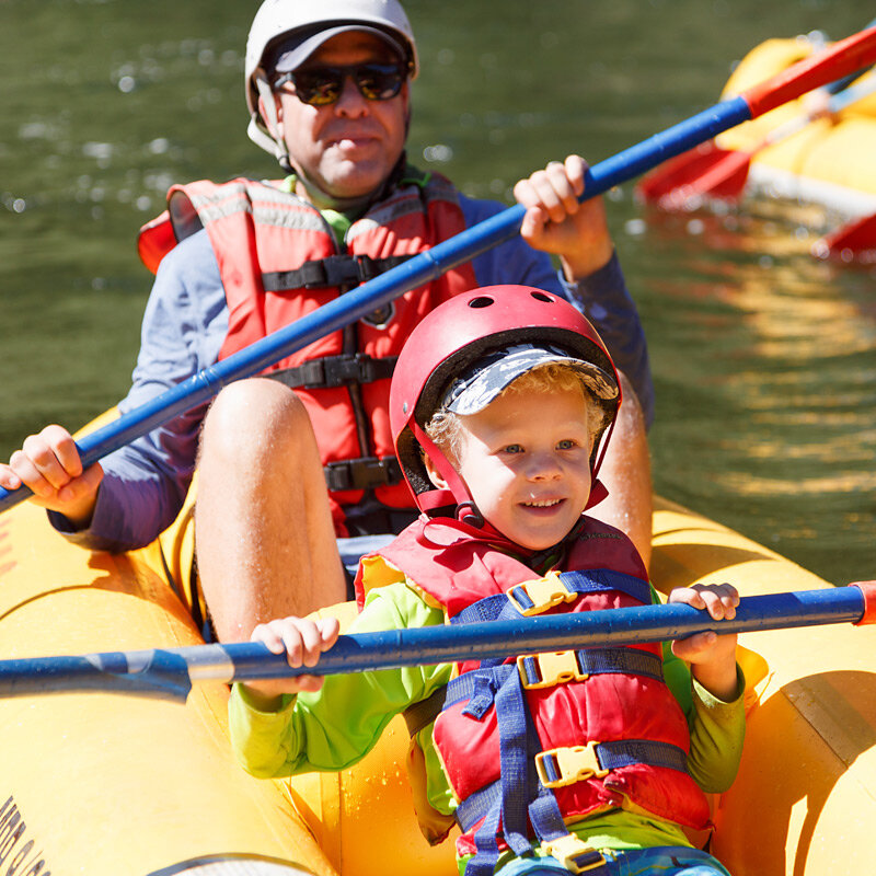 Close up of a young boy in the front of an inflatable kayak holding on to his paddle with an adult sitting in the seat behind him as they paddle down the Lower Klamath River.