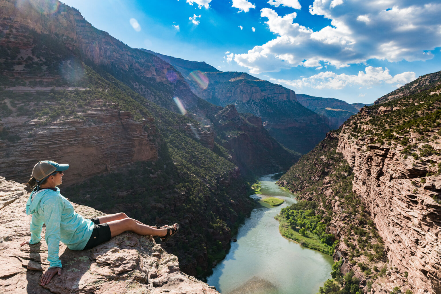 A woman sits towards the edge of the rim of a canyon wall and looks down at the Green River through the verdant Gates of Lodore.