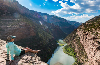 A woman sits towards the edge of the rim of a canyon wall and looks down at the Green River through the verdant Gates of Lodore.