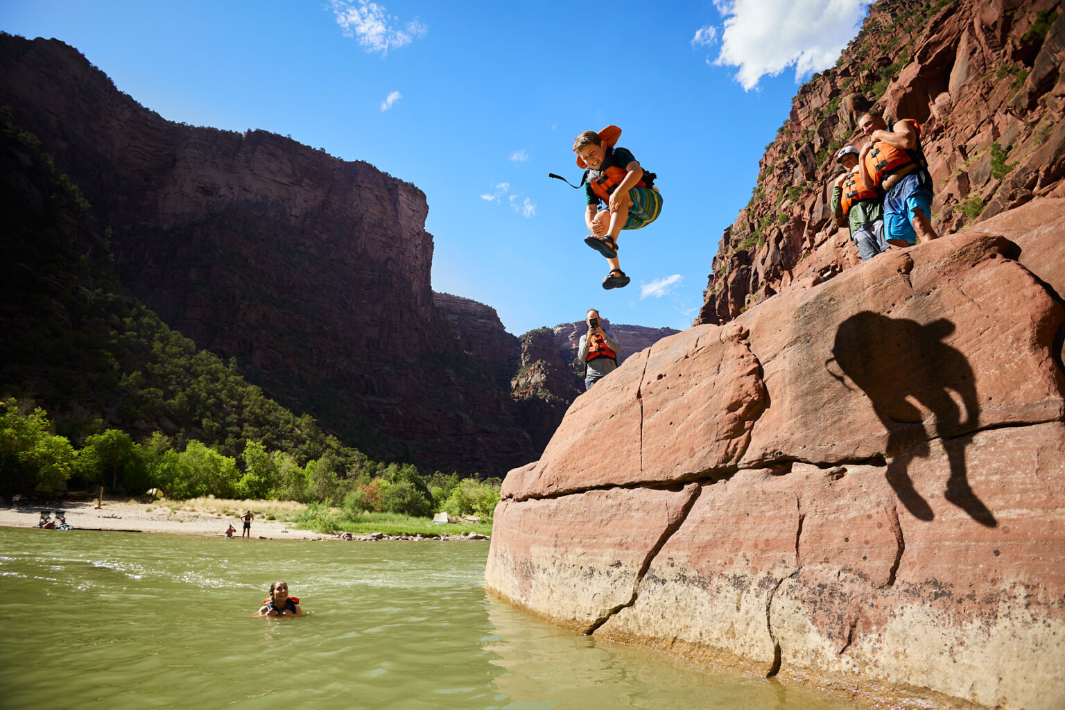 A boy mid-jump off a rock into the river as other guest waiting to jump watch and one guest who has already jumped in watches and smiles from the river.