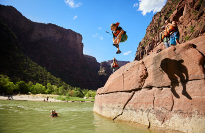 A boy mid-jump off a rock into the river as other guest waiting to jump watch and one guest who has already jumped in watches and smiles from the river.
