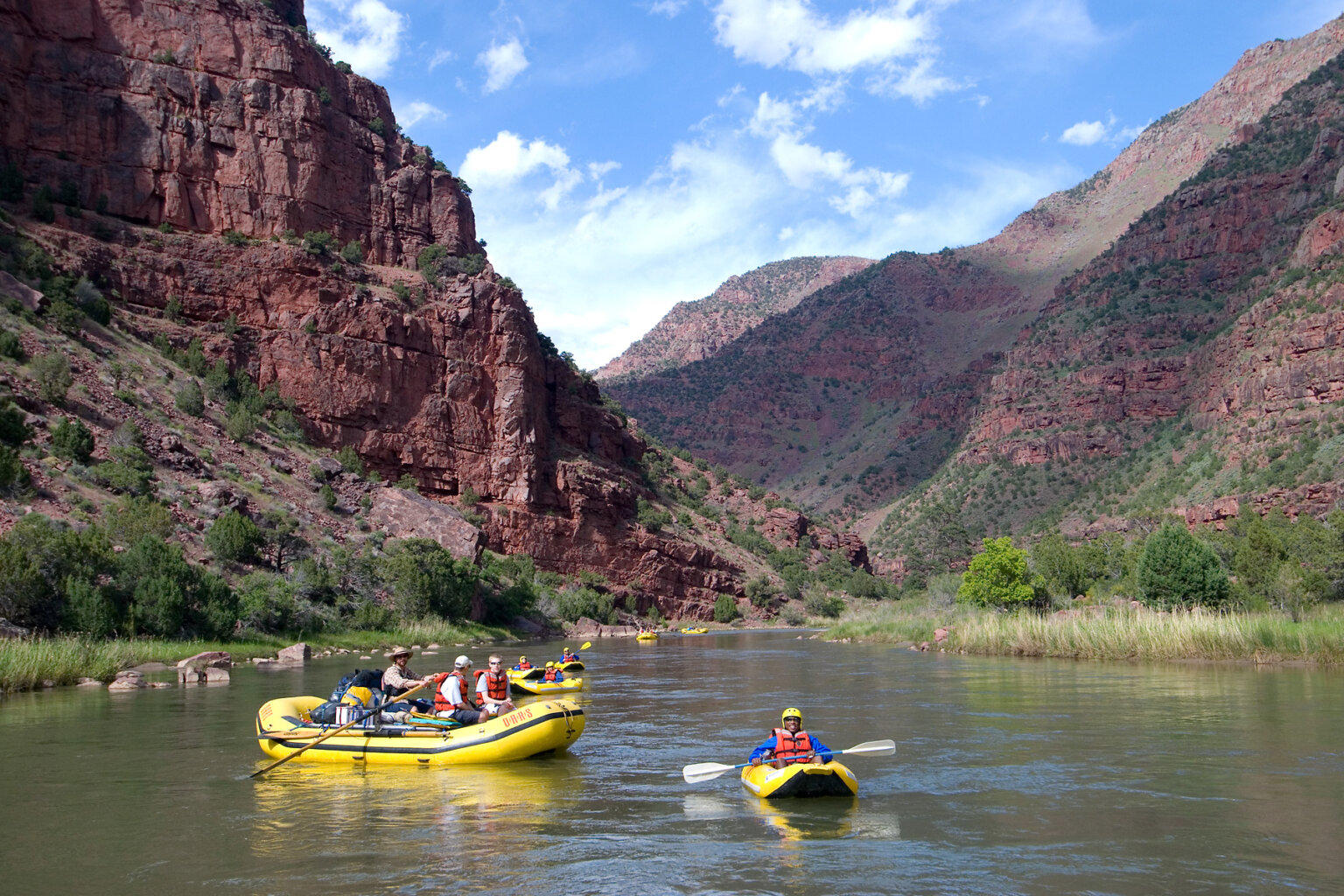 A gentleman in an inflatable kayak on the Green River smiles at the camera as several other kayakers and oar boats make their way downstream behind him, all surrounded by vibrant canyon walls studded with lots of green vegetation.