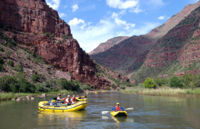 A gentleman in an inflatable kayak on the Green River smiles at the camera as several other kayakers and oar boats make their way downstream behind him, all surrounded by vibrant canyon walls studded with lots of green vegetation.