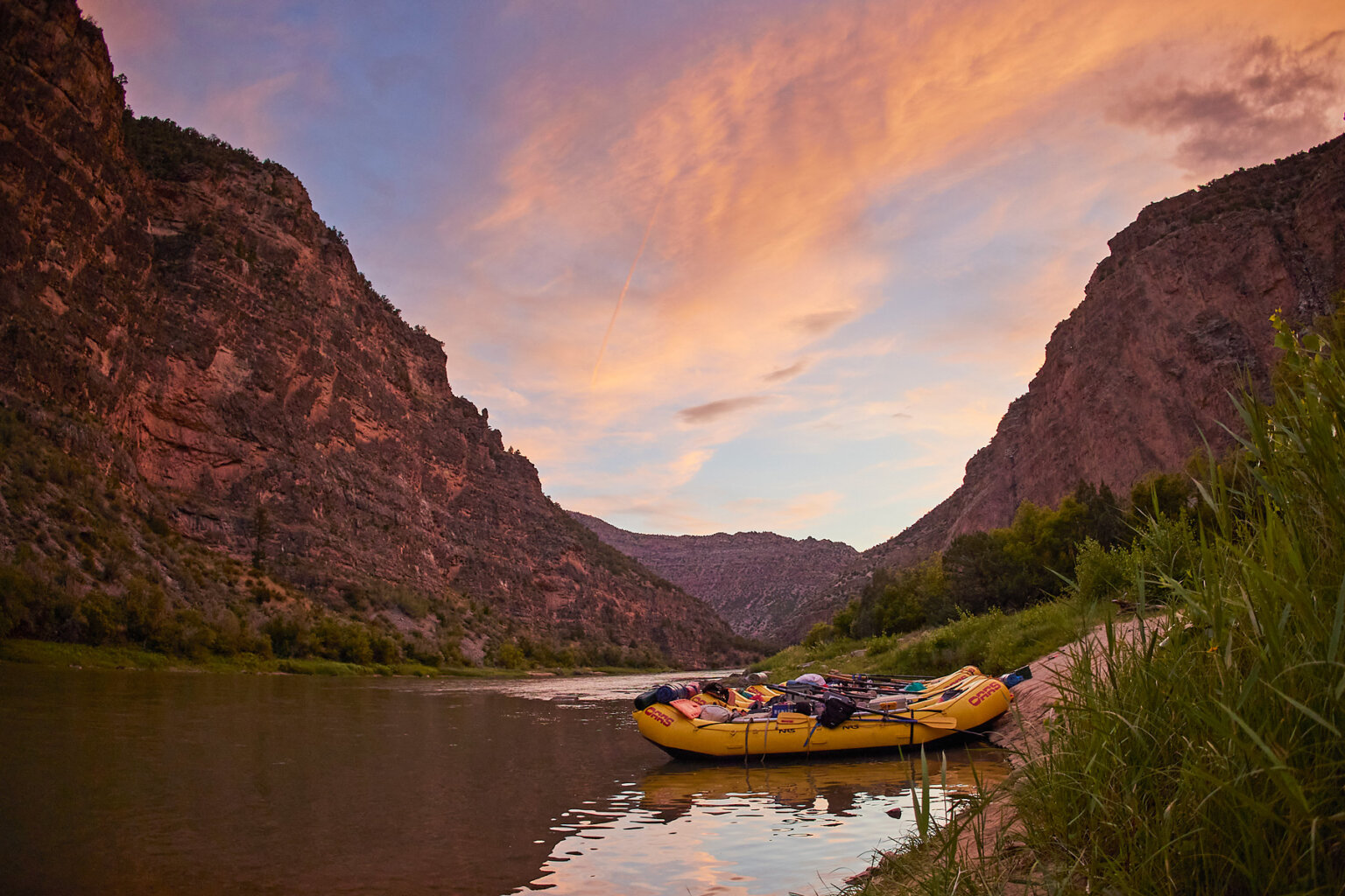 A glorious sunset overlooks several yellow rafts tied up on the shoreline of the Green River as vibrant red canyon walls surround.