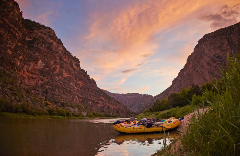 A glorious sunset overlooks several yellow rafts tied up on the shoreline of the Green River as vibrant red canyon walls surround.