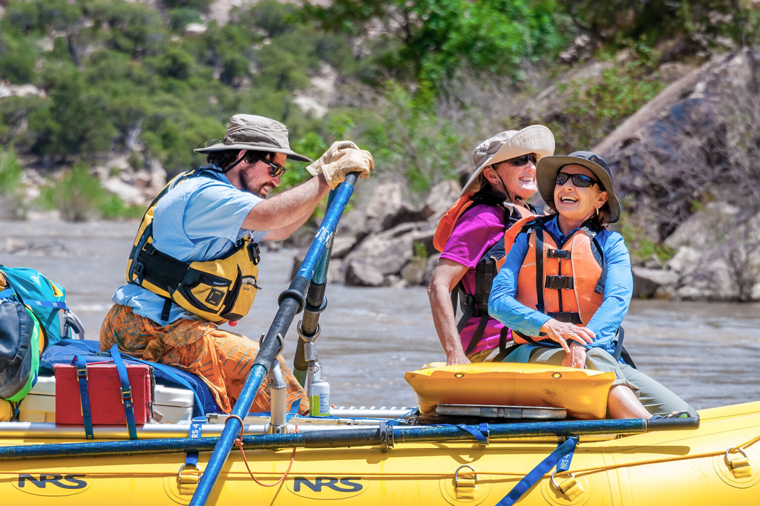 Sideview of two women sitting in the front of a raft smiling as they look up at the scenery and a guide rows.
