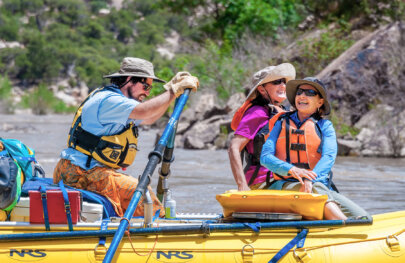 Sideview of two women sitting in the front of a raft smiling as they look up at the scenery and a guide rows.
