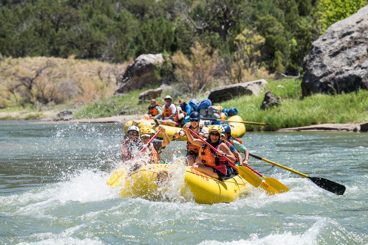 A raft full of paddlers splashes through a rapid as an oar raft with lounging guests wait behind them to make their run.