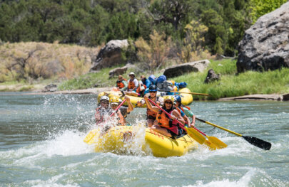 A raft full of paddlers splashes through a rapid as an oar raft with lounging guests wait behind them to make their run.