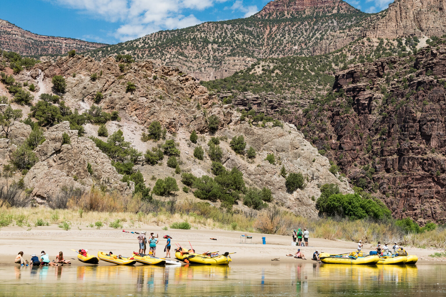 Several yellow rafts and inflatable kayaks pulled up on the shoreline while guests are scattered across the beach while guides prepare lunch.
