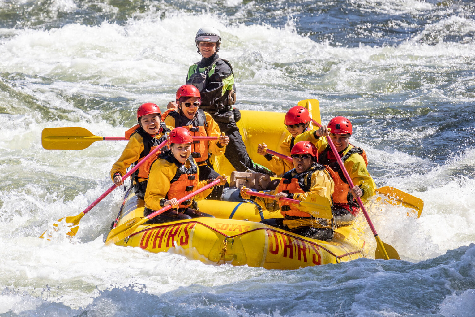 A raft full of ecstatic paddlers and a guide as they row through a stretch of frothy whitewater.