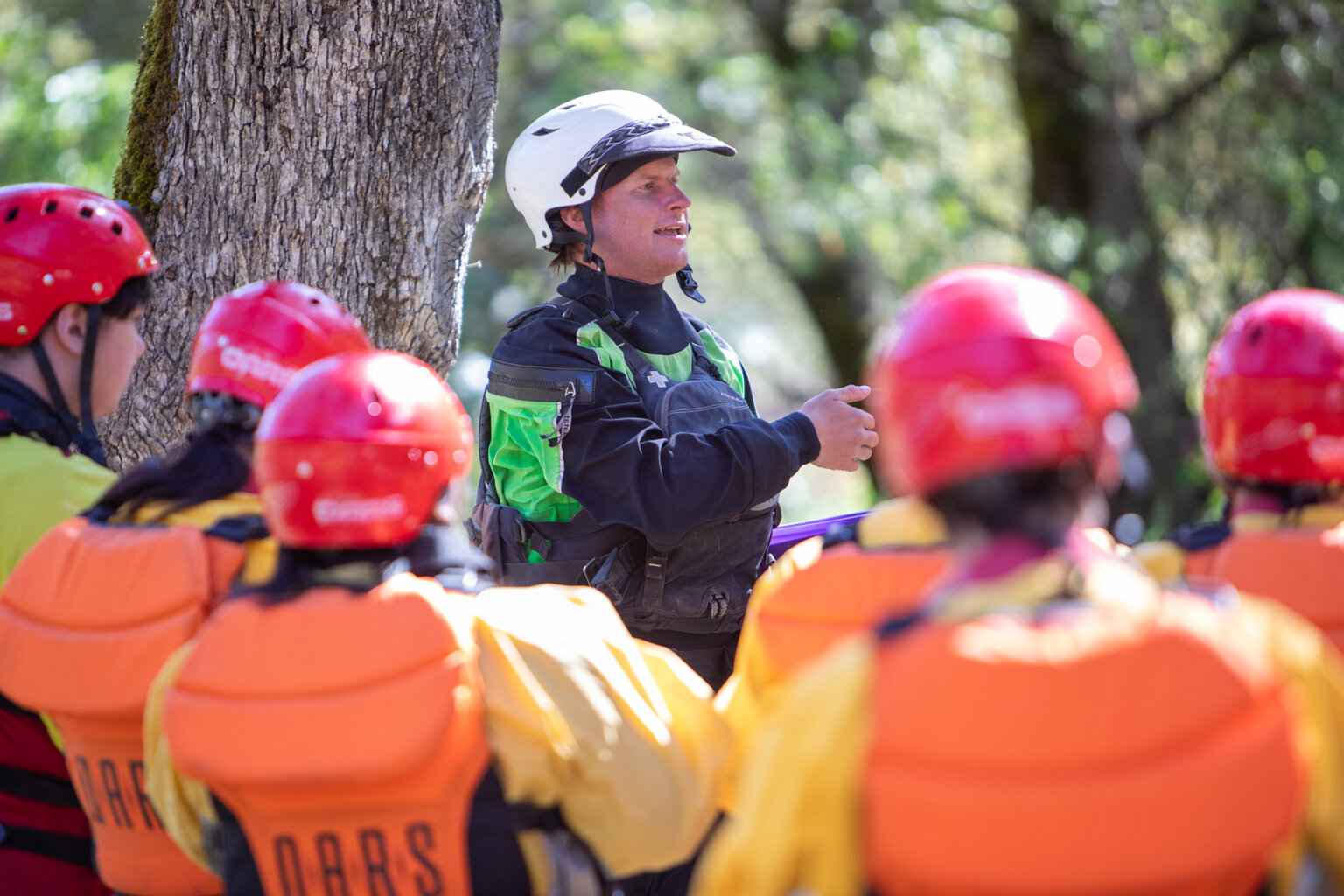 A guide stands in front of a tree and gives a safety talk as fully geared up rafters listen.