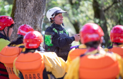 A guide stands in front of a tree and gives a safety talk as fully geared up rafters listen.