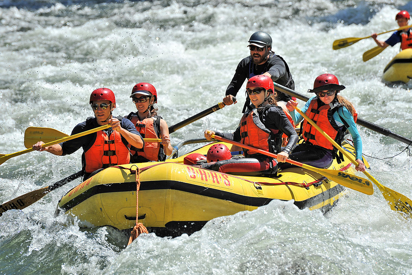 Four paddlers in a yellow raft with a guide rowing on oars as they paddle through frothy whitewater on the Merced River.